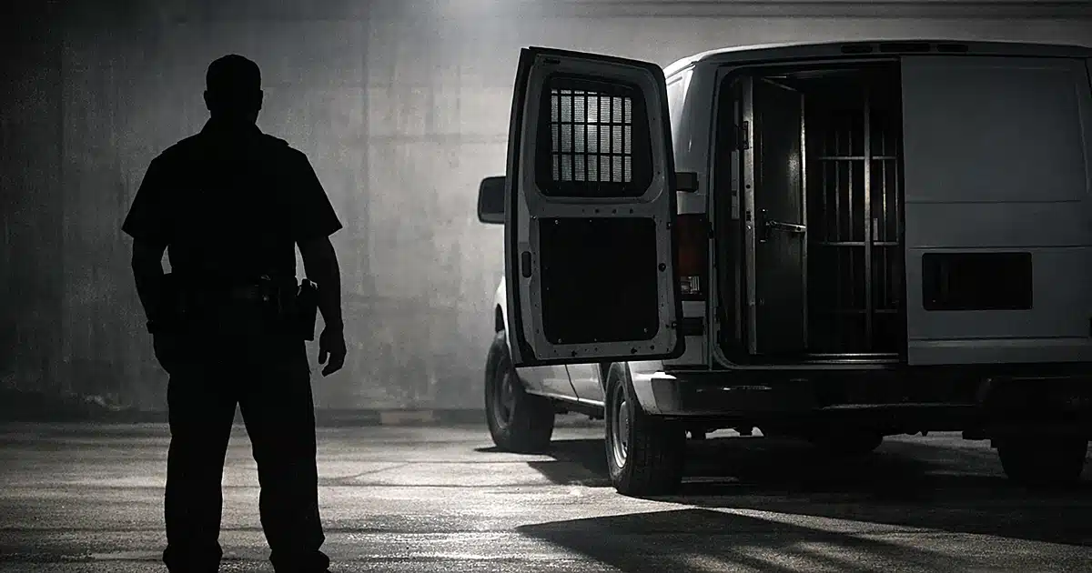 Silhouette of deputy beside transport van with barred windows in empty lot under harsh lighting