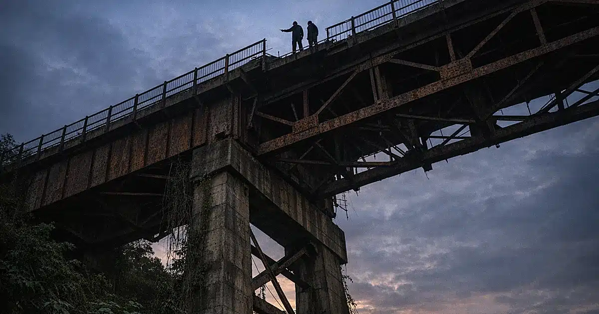 Abandoned bridge structure shot from below showing rusted steel beams and railings against darkening sky