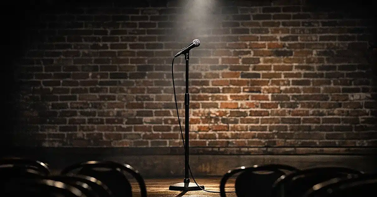Empty comedy club stage with microphone stand under spotlight and silhouetted chairs in foreground