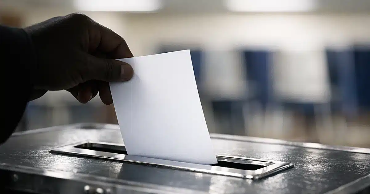 Silhouetted hand inserting blank ballot into metal ballot box slot at polling station