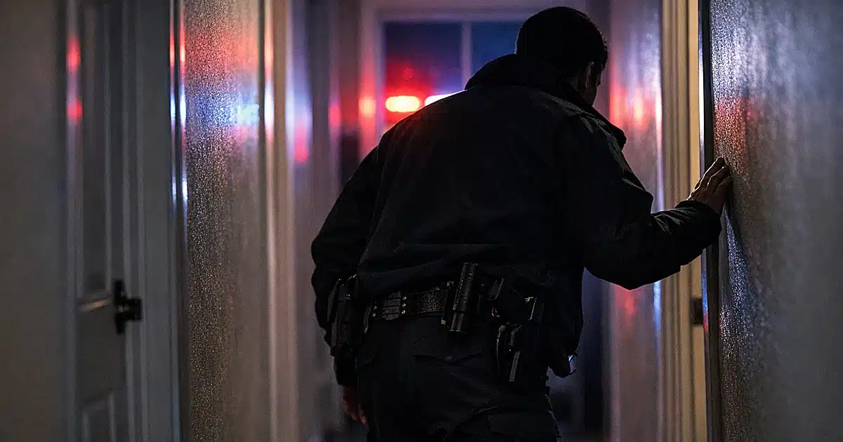 Police detective silhouette examining doorway in residential hallway with emergency lights flashing through window