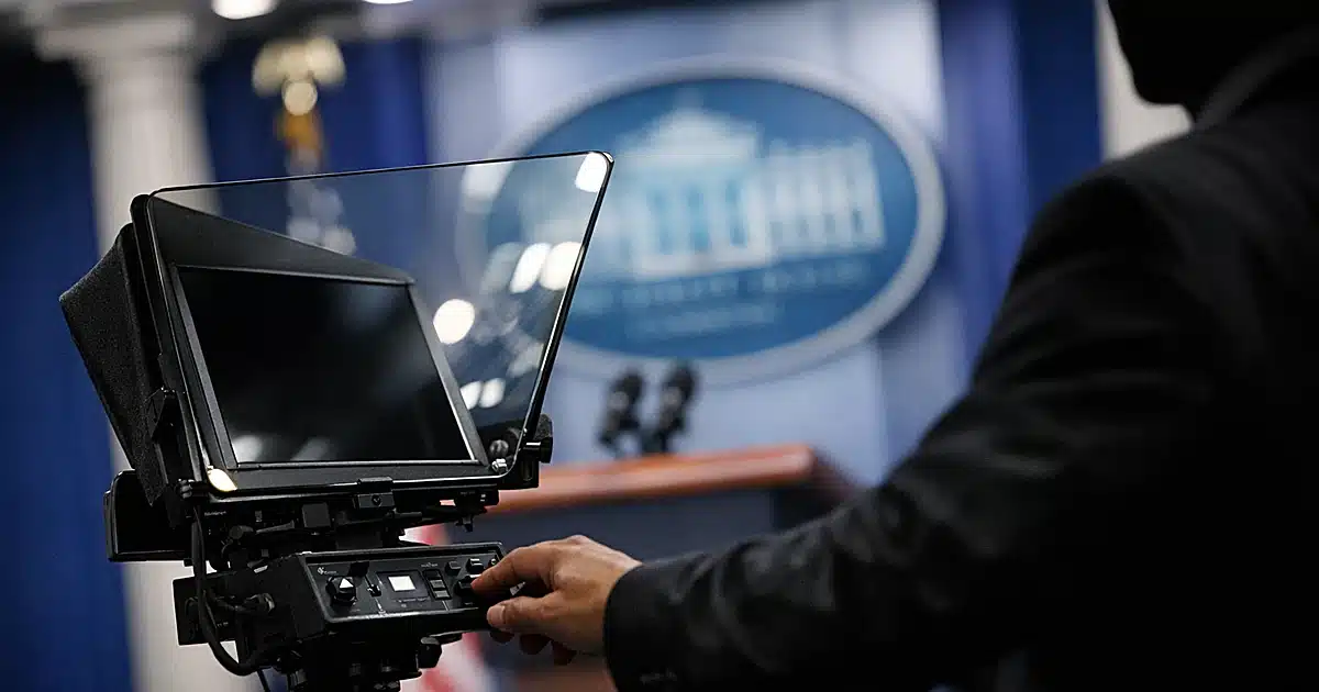 Presidential teleprompter screen on stand in White House-style briefing room with silhouetted figure reaching toward it