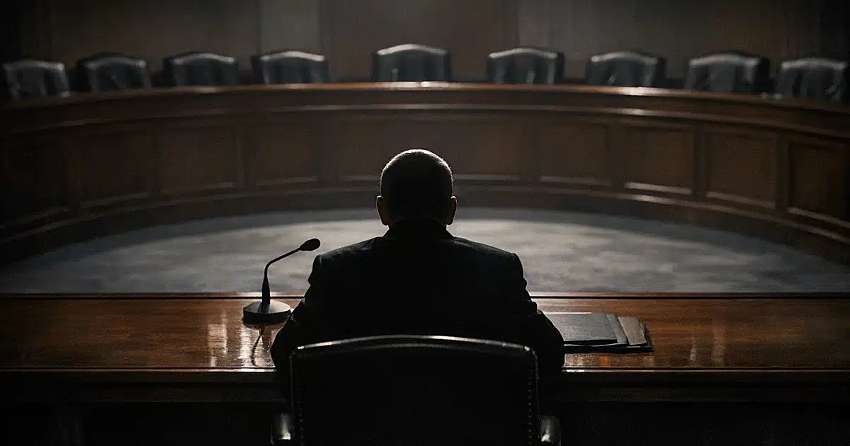 Silhouetted figure at witness table facing empty semicircular arrangement of chairs in congressional hearing room