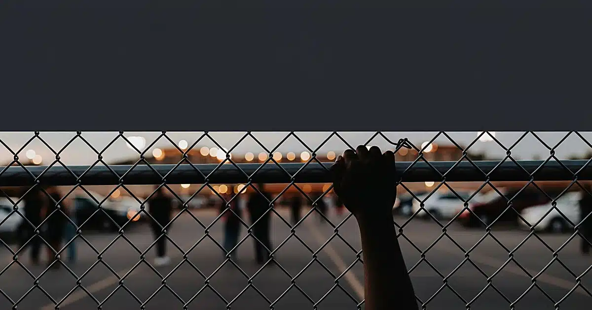Close-up of chain-link fence with hand gripping wire, blurred concert venue lights in background at dusk