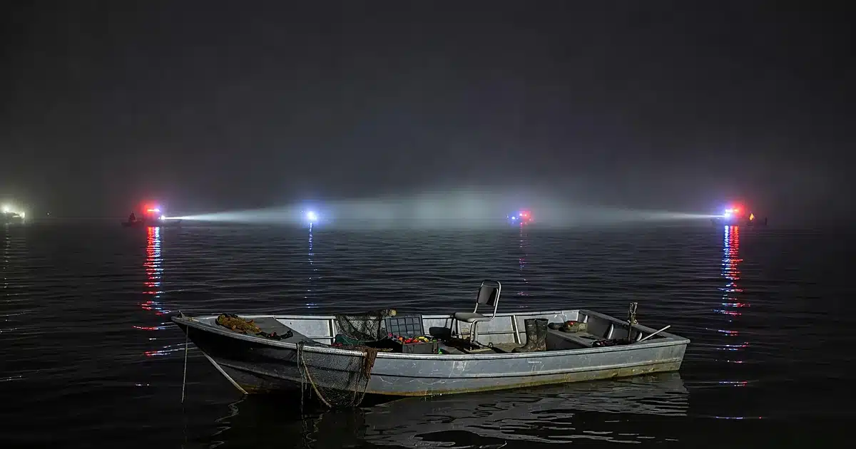 Empty fishing boat illuminated by searchlight on dark foggy water at night with emergency lights reflecting
