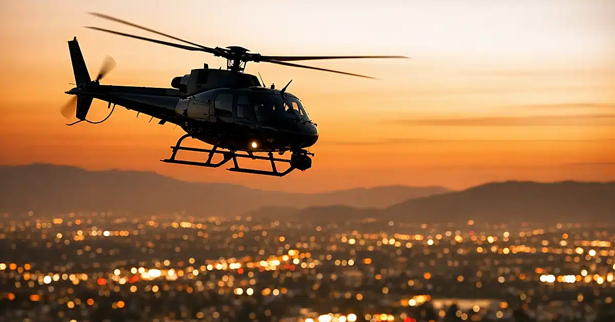 News helicopter silhouetted against orange dusk sky over blurred Los Angeles cityscape below