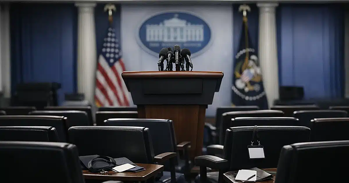 Empty White House press briefing room with scattered credentials on chairs facing podium with microphones