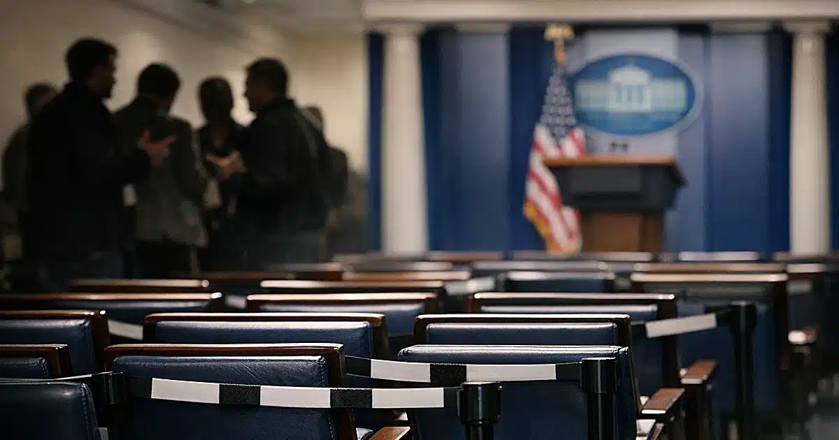 Empty press briefing room with cordoned-off seats and silhouetted reporters gathered to the side