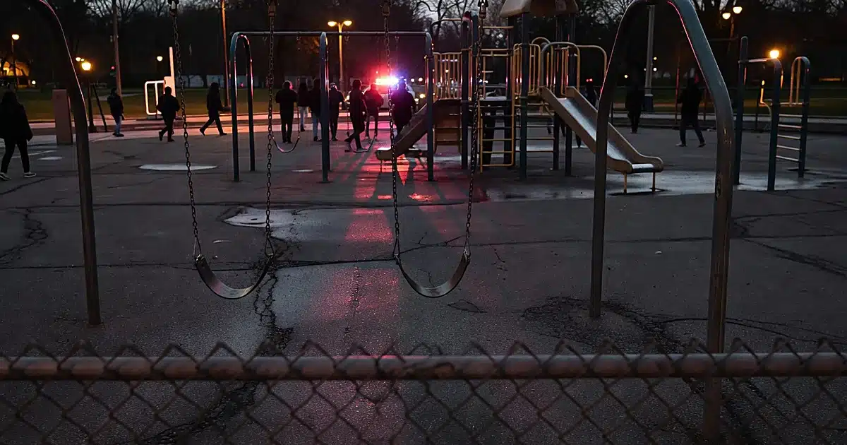 Urban playground at dusk with emergency lights reflecting off wet pavement and scattered silhouettes in background
