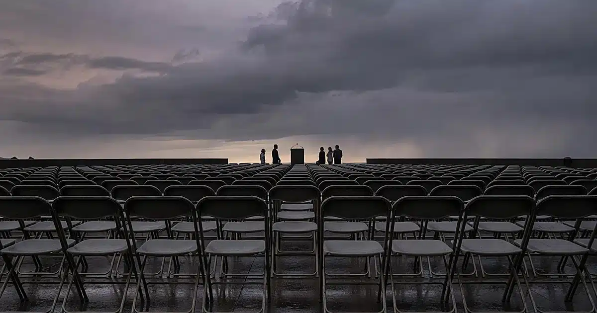 Empty memorial ceremony chairs face distant podium silhouettes at dusk with overcast sky
