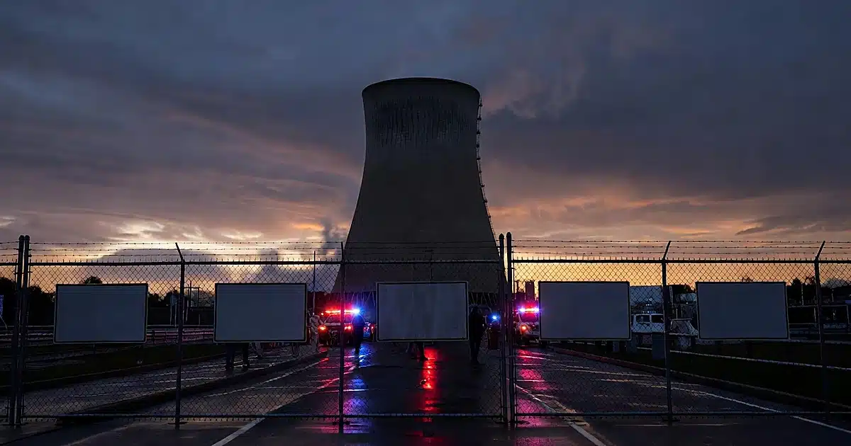 Nuclear power plant cooling tower behind security fence with emergency vehicle lights reflecting on wet pavement at dusk