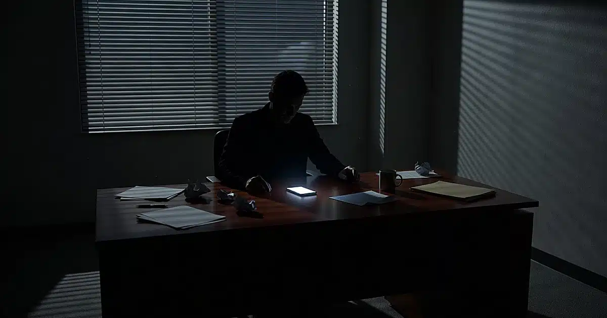 Silhouetted figure at office desk with glowing smartphone in darkened corporate workspace with venetian blind shadows