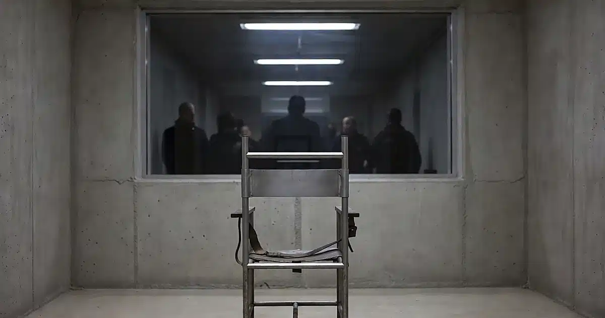 Military execution chamber with metal chair facing observation window, silhouettes visible beyond glass