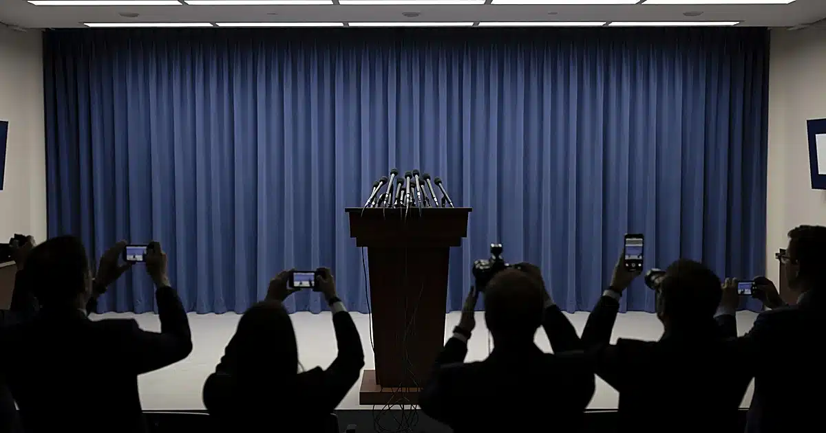 Empty White House press briefing room podium with microphones, reporters visible as silhouettes from behind