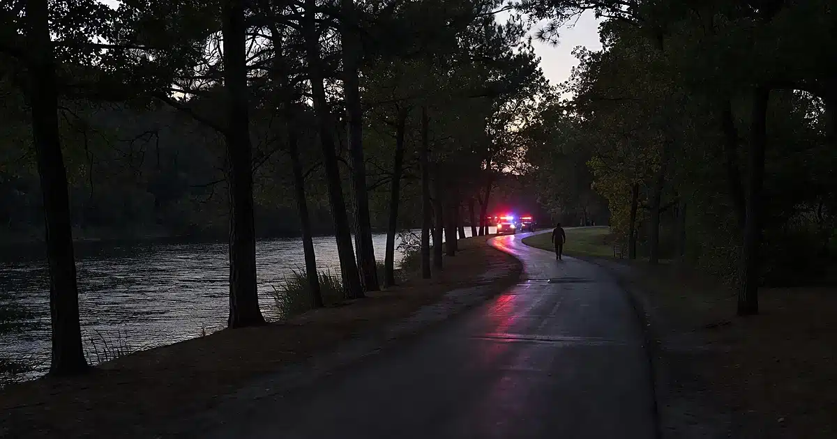 Empty riverside trail at dusk with emergency lights reflecting on wet pavement and distant silhouetted figure