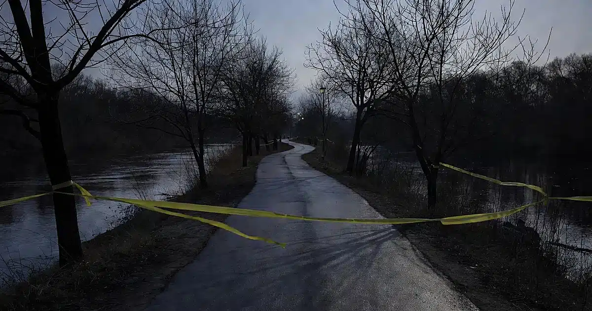 Empty river trail at dusk with crime scene tape across entrance, bare trees and dark water visible