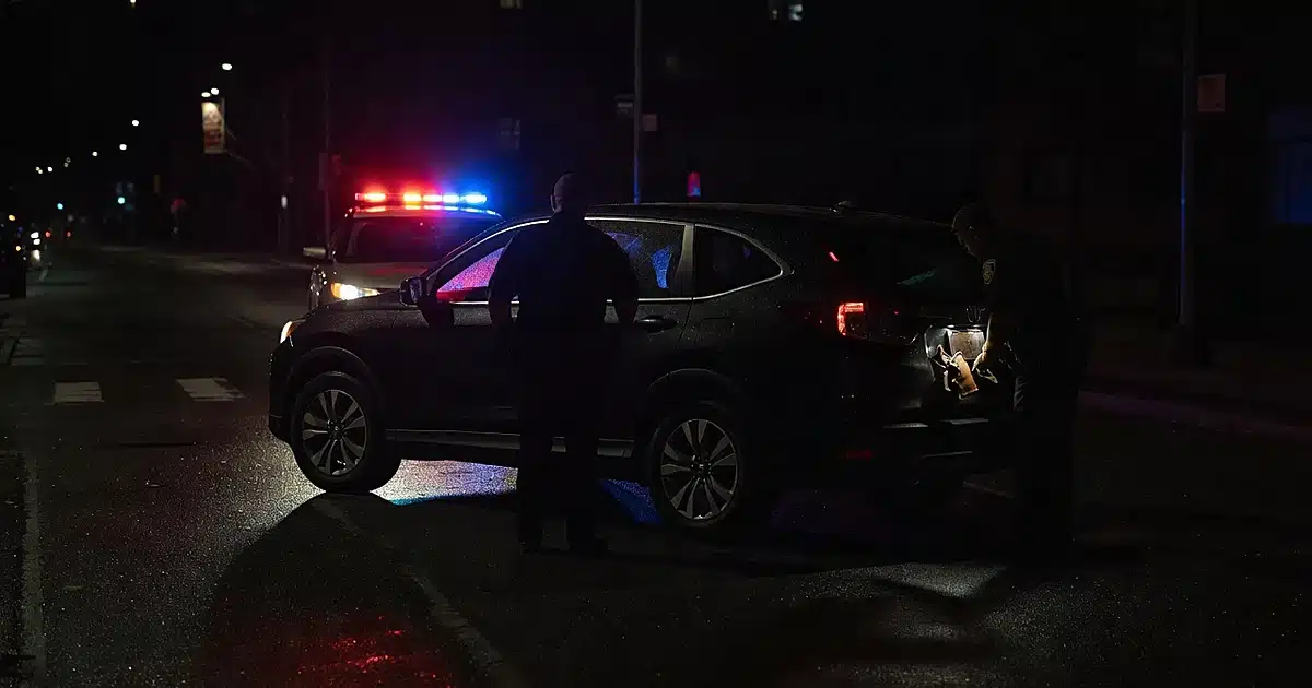 Traffic stop at night with SUV, emergency lights, and silhouetted officers examining vehicle with holster visible