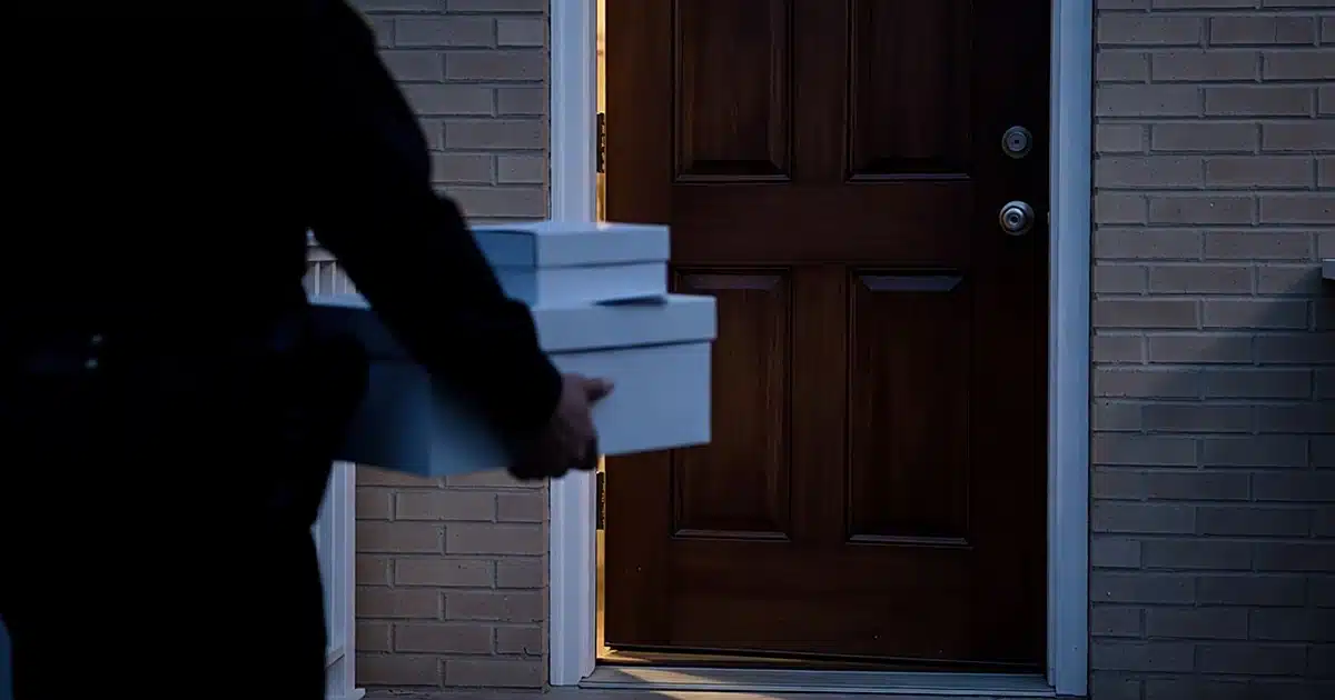 Pre-dawn federal raid scene showing silhouetted agent with document boxes at residential doorway
