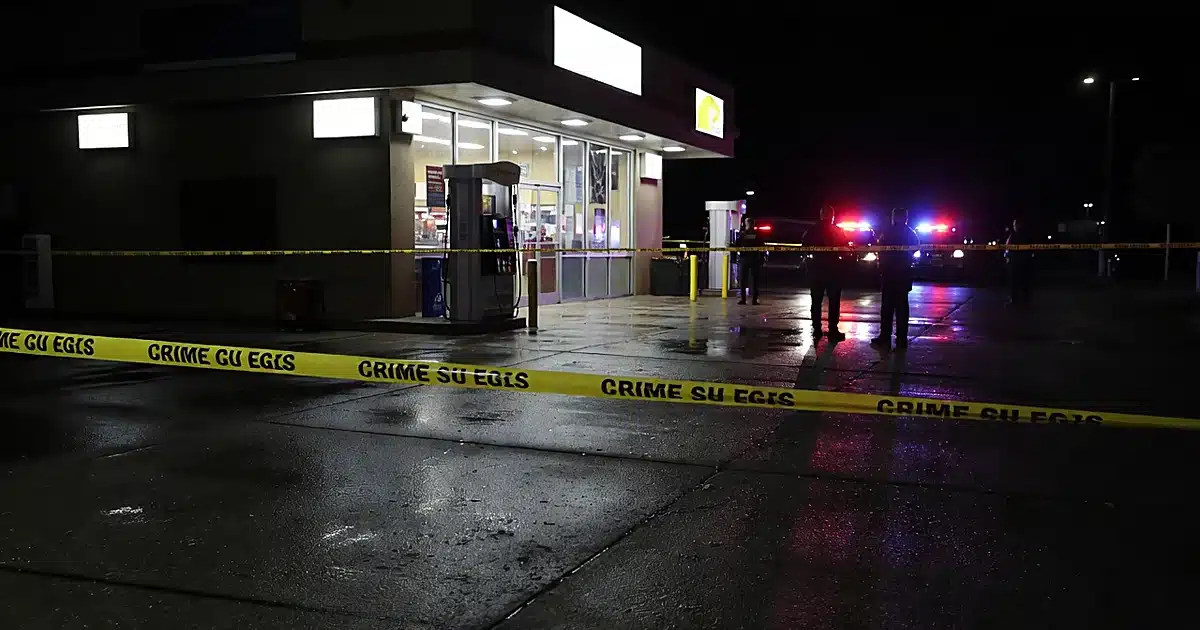 Crime scene tape at gas station entrance with police vehicle lights reflecting on wet pavement at night